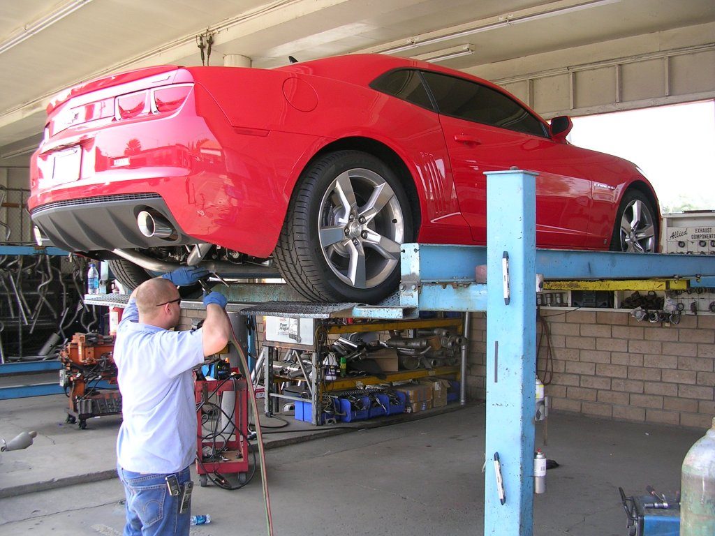 A man is repairing a red car inside a well-lit garage, surrounded by tools and equipment.